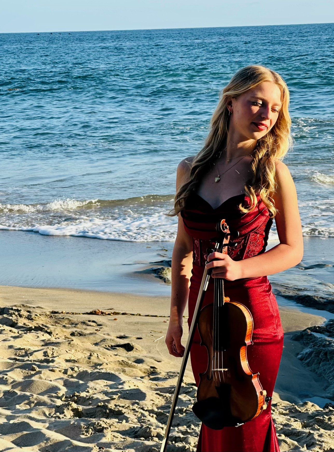 Hannah in a red gown on the beach, holding her violin
