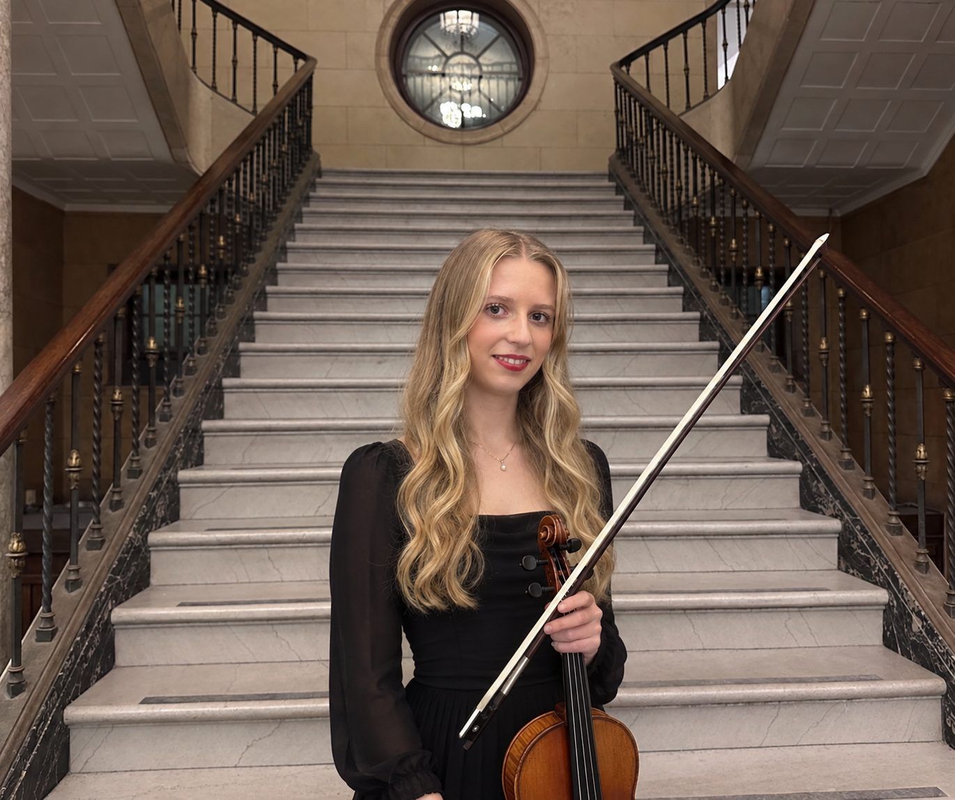 Hannah in black with her violin at the foot of a marble staircase